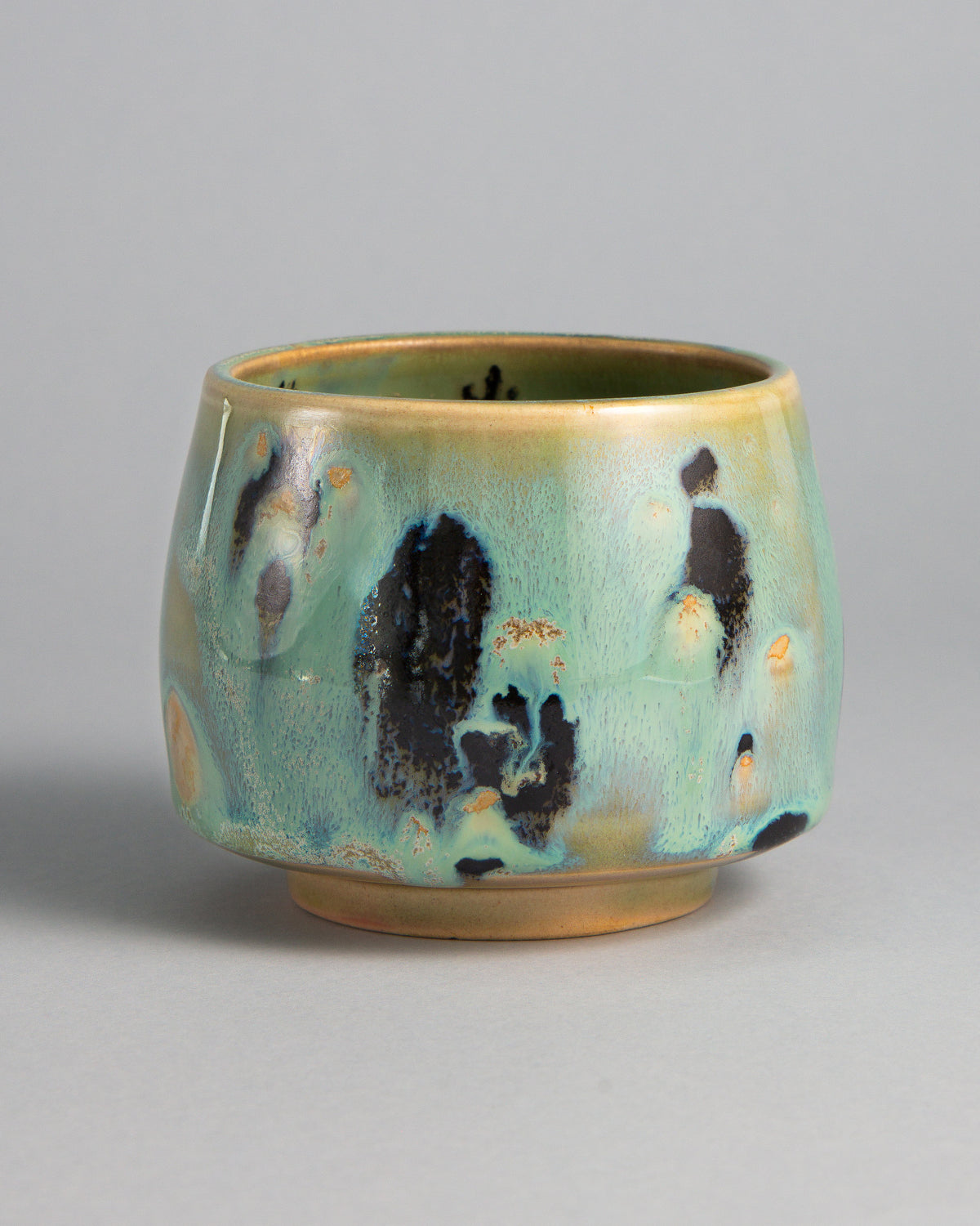 WINDY CUP 'LICHEN', handmade stoneware cup without a handle by Studio 517. Glossy seafoam green with large black spots edged in blue and small amber flecks, fading to sandy beige at the rim and foot, like lichen growing on stone.