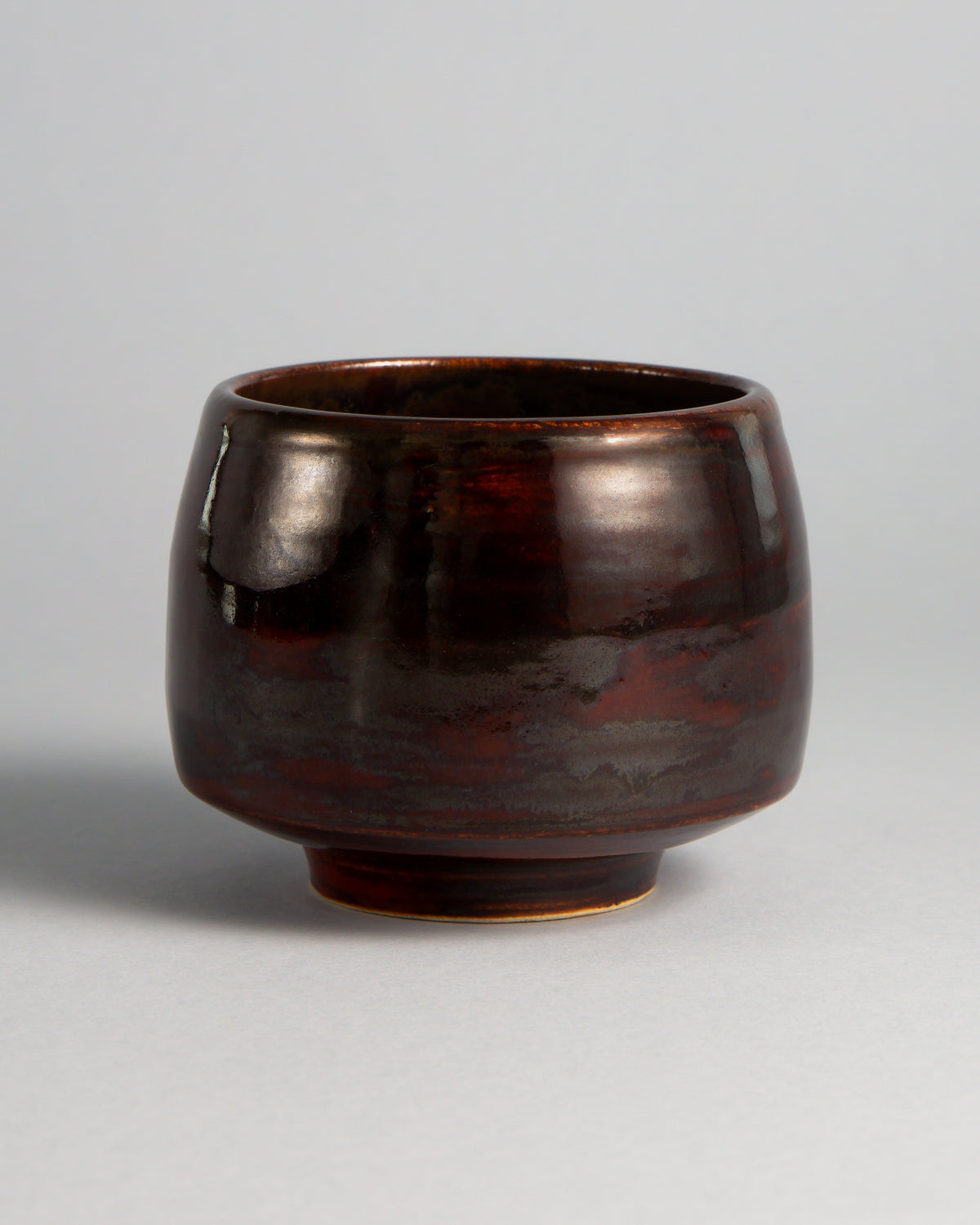 WINDY CUP 'MAGMA', taza de gres sin asa hecha a mano por Studio 517. Bronce oscuro muy brillante, casi negro, con bandas de rojo intenso que asoman en la mitad inferior, como lava enfriada que aún guarda el calor.