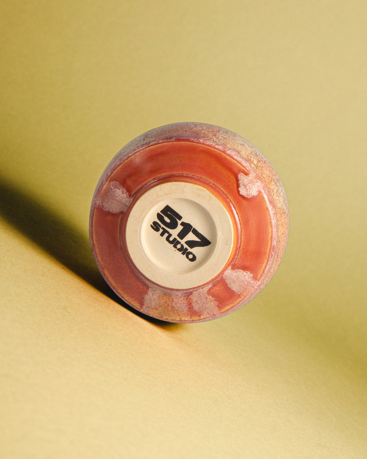 WINDY handmade stoneware cup by Studio 517 lying on its side on a yellow background, showing its base: rust pink glaze with white mottling around the unglazed foot stamped 517 STUDIO.