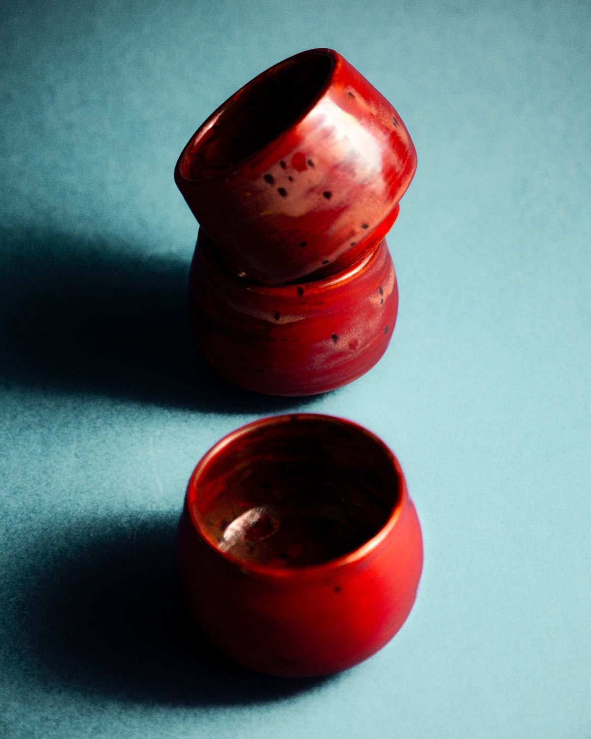 WINDY handmade stoneware cups by Studio 517: two glossy red cups stacked and tilted, a third upright in front, on a flat teal background with soft shadows. Small dark specks on the red glaze.