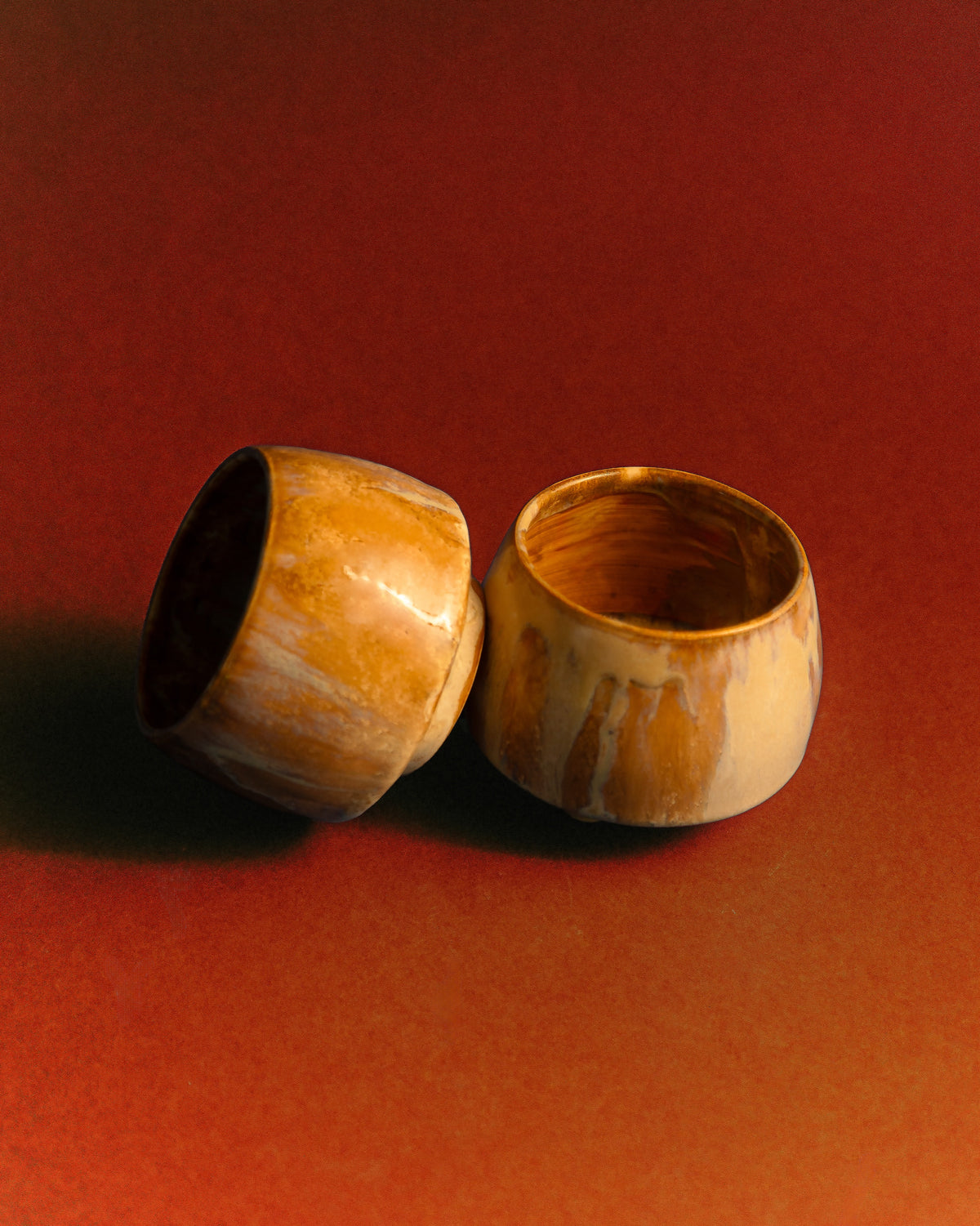 WINDY handmade stoneware cups by Studio 517: two cups side by side on a rust red background, one tipped on its side. Cream glaze with caramel and brown streaks running down.