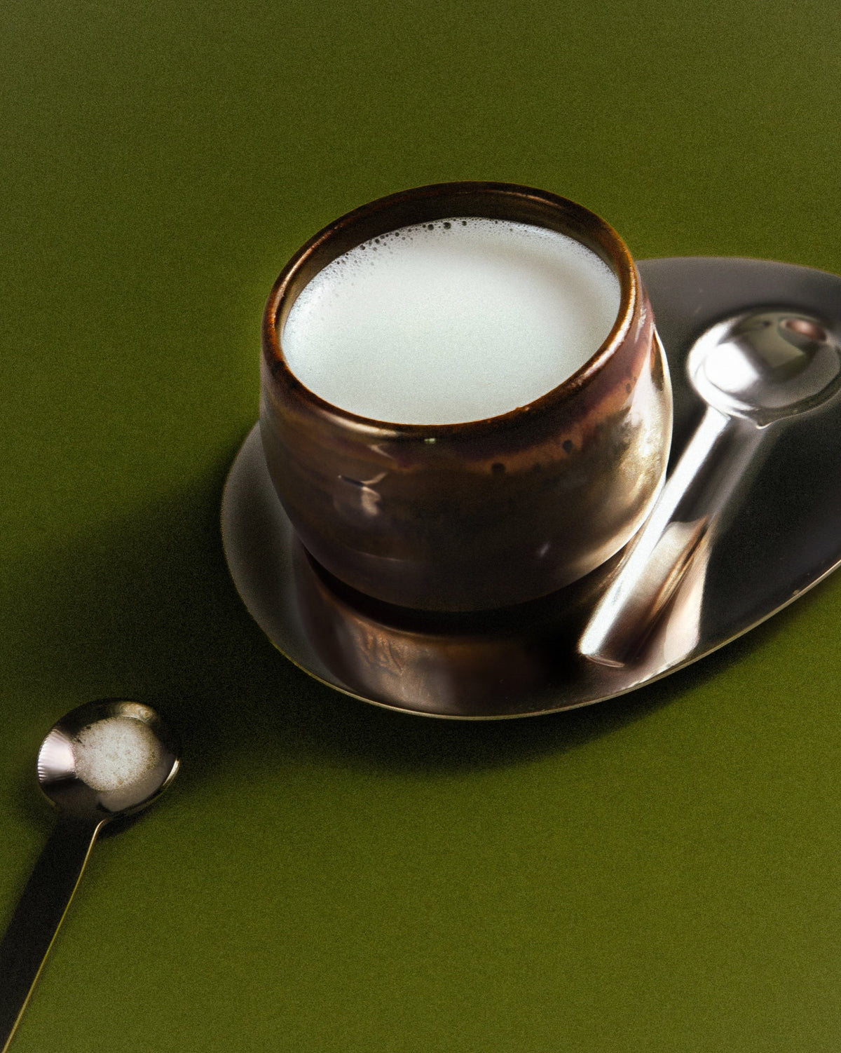 WINDY handmade stoneware cup by Studio 517 in glossy dark brown, filled with milky coffee, on a dark oval saucer with a silver spoon. A second spoon lies beside it on an olive green background.