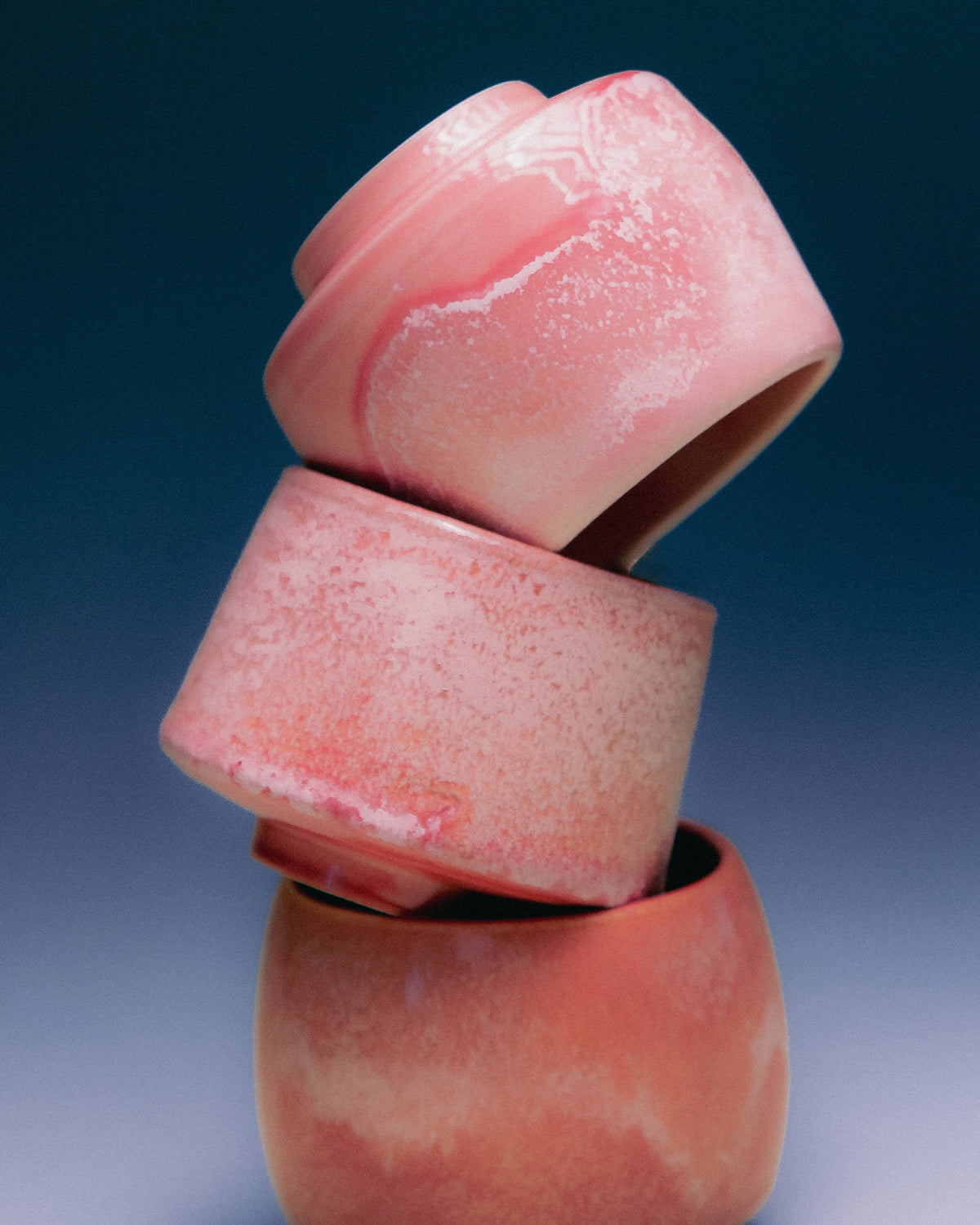 WINDY handmade stoneware cups by Studio 517: three pink cups stacked at angles against a dark blue background that fades to grey. Satin pink glaze with white mottled patches.