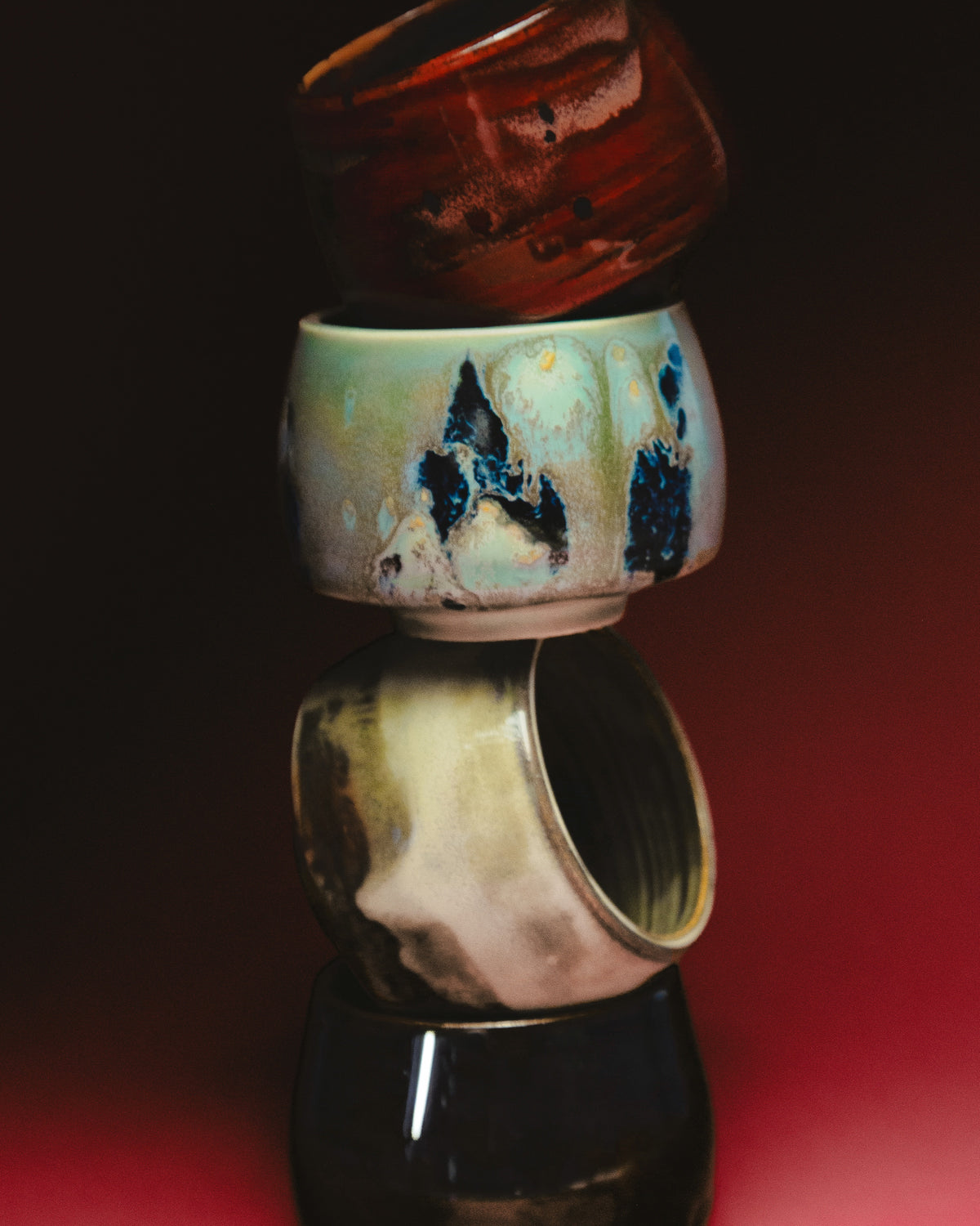 WINDY handmade stoneware cups by Studio 517: four cups stacked in a tall tower on a black to red background. Glossy red, white with dark blue drips, cream with brown streaks, and black.