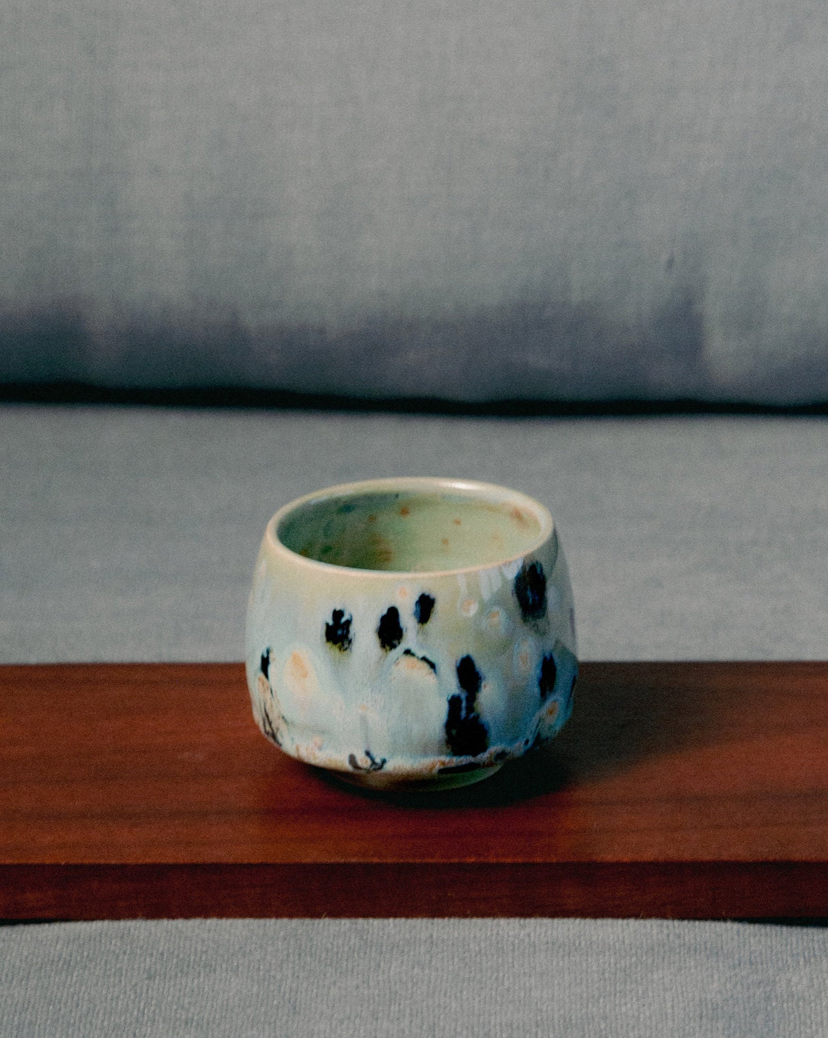 WINDY handmade stoneware cup by Studio 517 on a wooden bench in front of a grey sofa. White glaze with irregular dark blue and black spots, pale green inside.